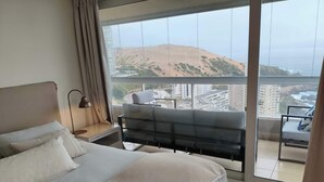 A hotel room with a large bed, a desk with a lamp, a chair, and a balcony with a view of the city and hills. - Modern 4BR Apartment with Ocean View near Concón (Valparaíso)