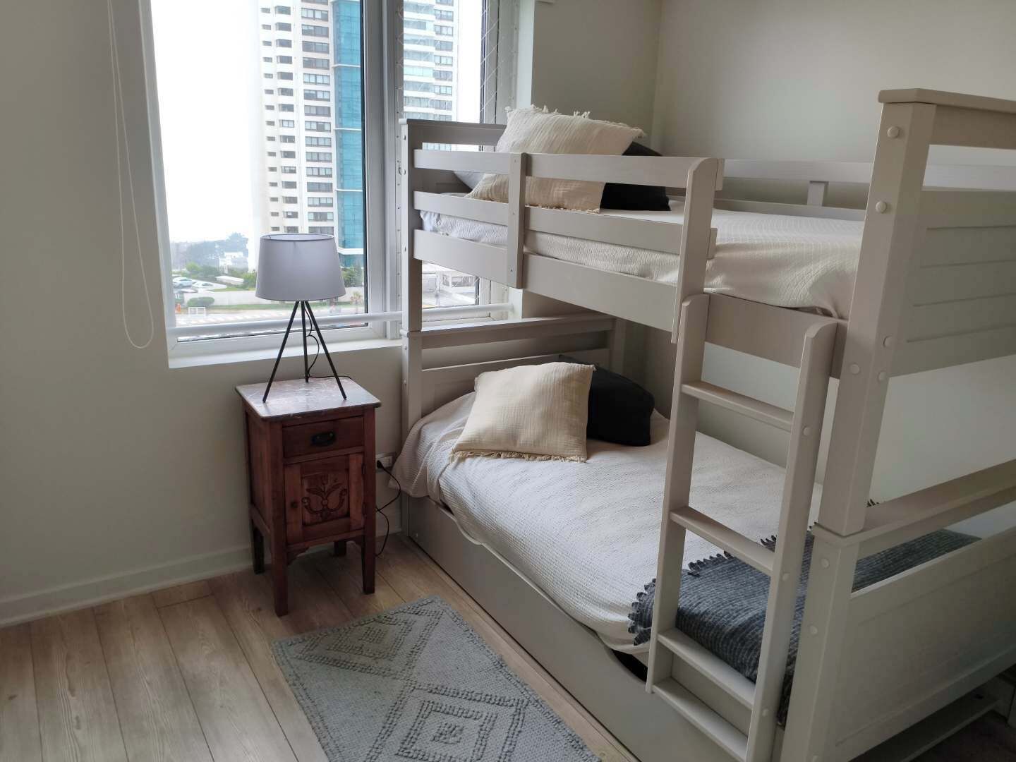 Une chambre avec un lit superposé, une petite table de chevet en bois, une lampe et une vue sur de hauts immeubles par la fenêtre.