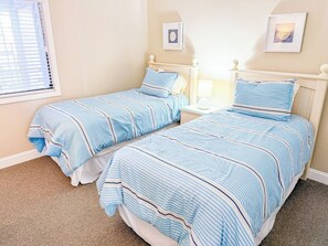 A bedroom with two beds, each with striped bedding, a bedside table with a lamp, and a window with blinds. - Tilghman Beach and Racquet Club 321 (North Myrtle Beach)