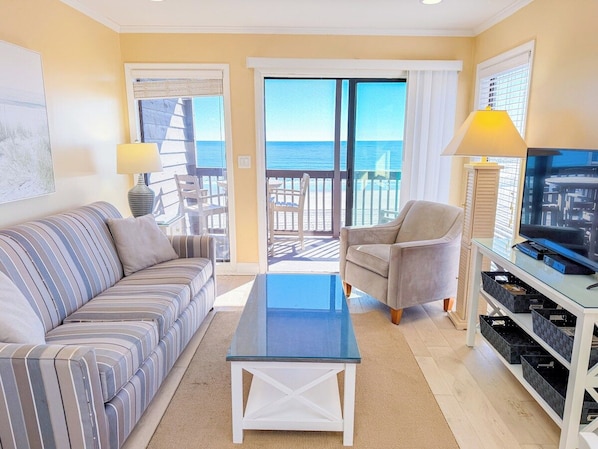 A living room with a sofa, armchair, coffee table, TV, and a view of the ocean through a sliding door.