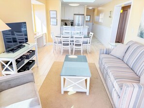 A living room with a sofa, a TV, a dining table with chairs, and a kitchen in the background. - Tilghman Beach and Racquet Club 321 (North Myrtle Beach)