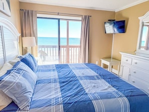 A bedroom with a bed, a television, a window with curtains, and a view of the sea.