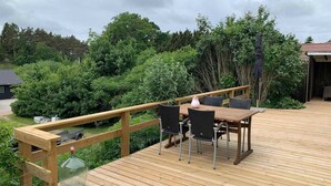 A wooden deck with a table and chairs, surrounded by lush greenery and trees. - 5 Star Holiday Home in Ebeltoft (Ebeltoft)