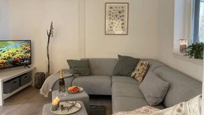 Ein modernes Wohnzimmer mit einem grauen Ecksofa, einem Flachbildfernseher, einem kleinen Tisch mit einer Kerze und einem gerahmten abstrakten Kunstwerk an der Wand.