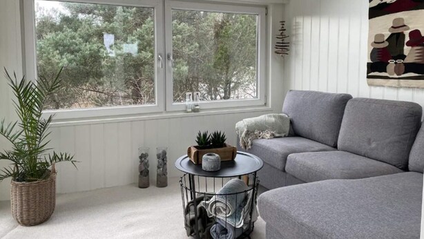 Ein gemütliches Wohnzimmer mit einem grauen Sofa, einem runden Tisch mit Pflanze und Korb, einem Blumentopf und einem Fenster mit Blick auf Bäume.