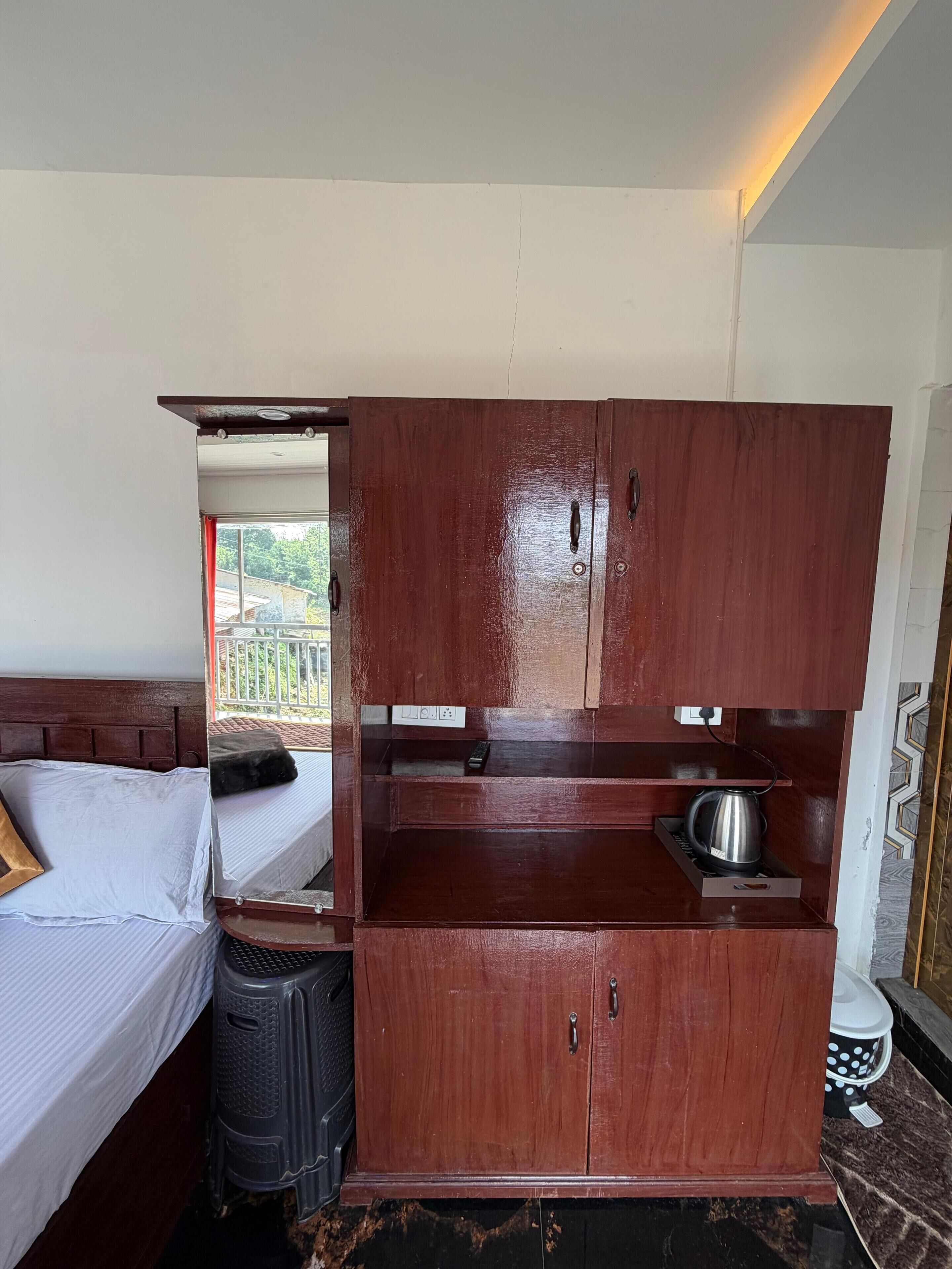 A compact room with a bed, a wooden wardrobe, a small kitchen area with a kettle, and a view of the outdoors.