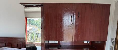 A compact room with a bed, a wooden wardrobe, a small kitchen area with a kettle, and a view of the outdoors.