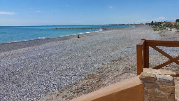 A pebbled beach with a wooden railing and stone wall, leading to a clear view of the sea.