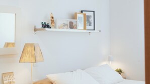A bedroom with a bed, bedside table, lamp, and wall-mounted shelf with decorative items.