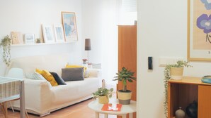 A living room with a white sofa, yellow and gray cushions, a small round table with a book and a plant, and a wall with framed pictures and a plant shelf.
