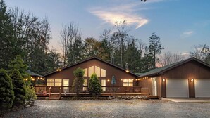 A two-story wooden cabin with a covered porch, large windows, and a gravel driveway.
