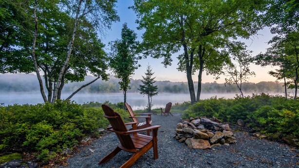 A serene lakeside setting with two Adirondack chairs, a pile of firewood, and a dense forest backdrop.