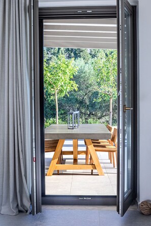 Une porte-fenêtre coulissante donne sur une terrasse extérieure où se trouvent une table et des chaises en bois, le tout entouré de verdure.