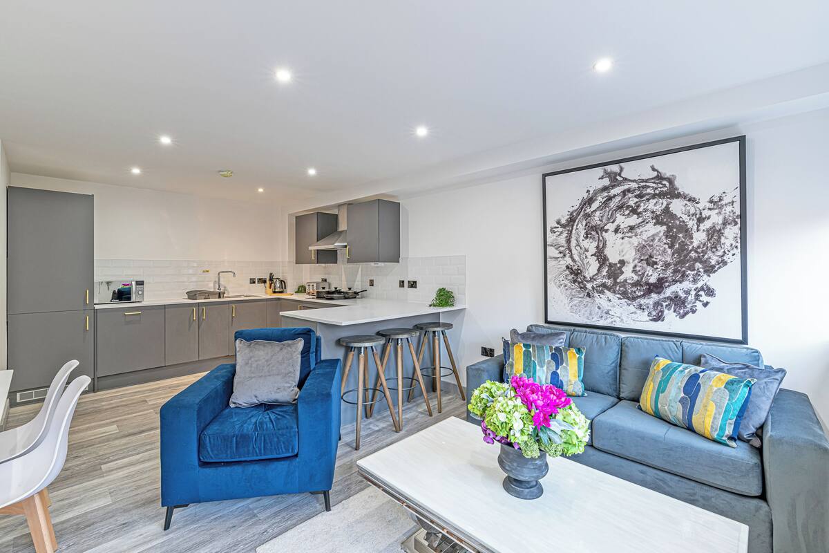 A modern living room with a blue sofa, a coffee table with flowers, and a kitchen in the background.