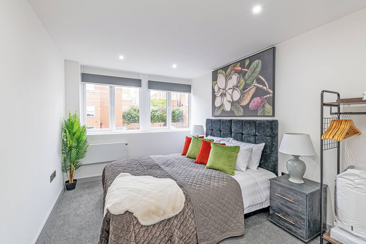 A bedroom with a bed, a nightstand, a lamp, a plant, and a window with a view of buildings.