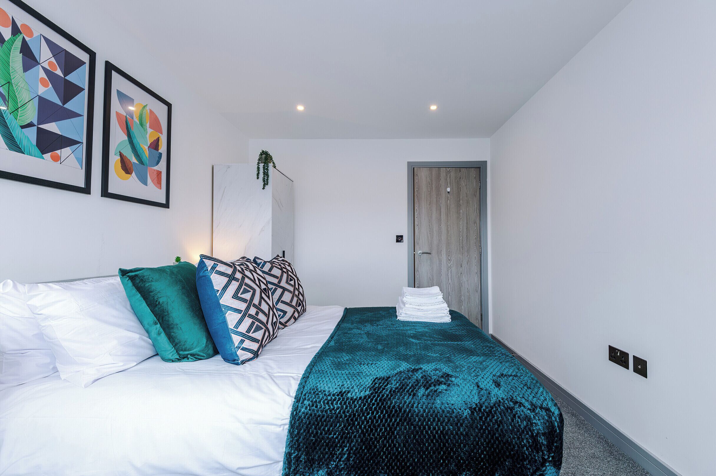 A neatly made bed with white linens, teal and black patterned pillows, and a teal blanket. There are two framed abstract artworks on the wall, a wooden door, and a small plant on the wall.