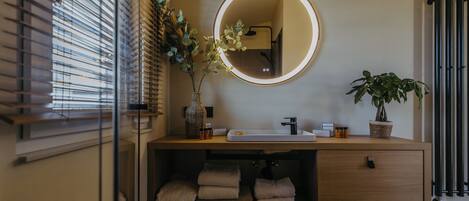 Une salle de bain avec un meuble-lavabo en bois, un miroir rond avec un éclairage intégré, un lavabo et deux plantes en pot.