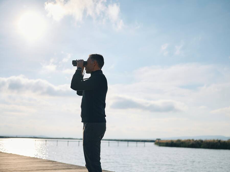 A person standing on a wooden pier, holding a camera, with a body of water and a bridge in the background.