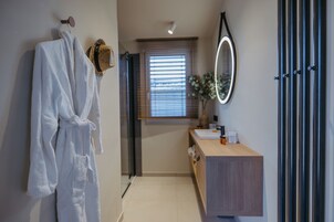 Un baño moderno con una bata blanca colgada en un gancho, un tocador de madera con un espejo y una ventana con persianas.