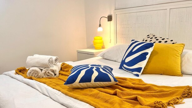 Ein ordentlich bezogenes Bett mit weißer Bettwäsche, einer senffarbenen Tagesdecke und zwei blau-weißen Kissen.
