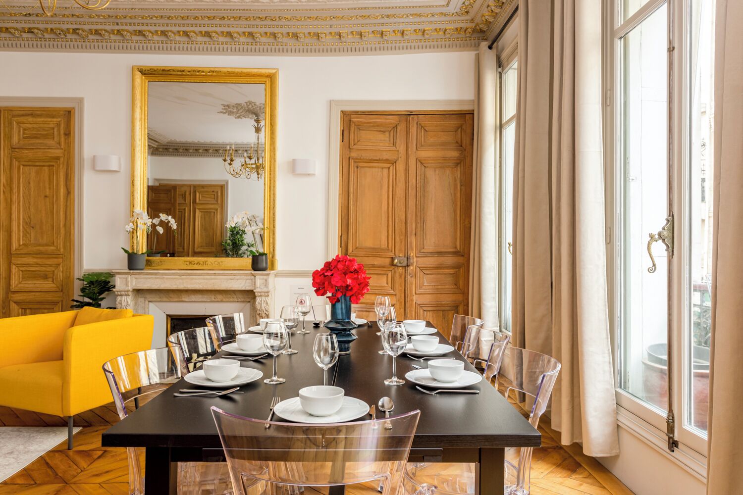 A dining room with a long table set for six, surrounded by clear chairs, with a yellow sofa and a fireplace in the background.