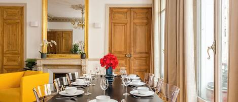 A dining room with a long table set for six, surrounded by clear chairs, with a yellow sofa and a fireplace in the background.