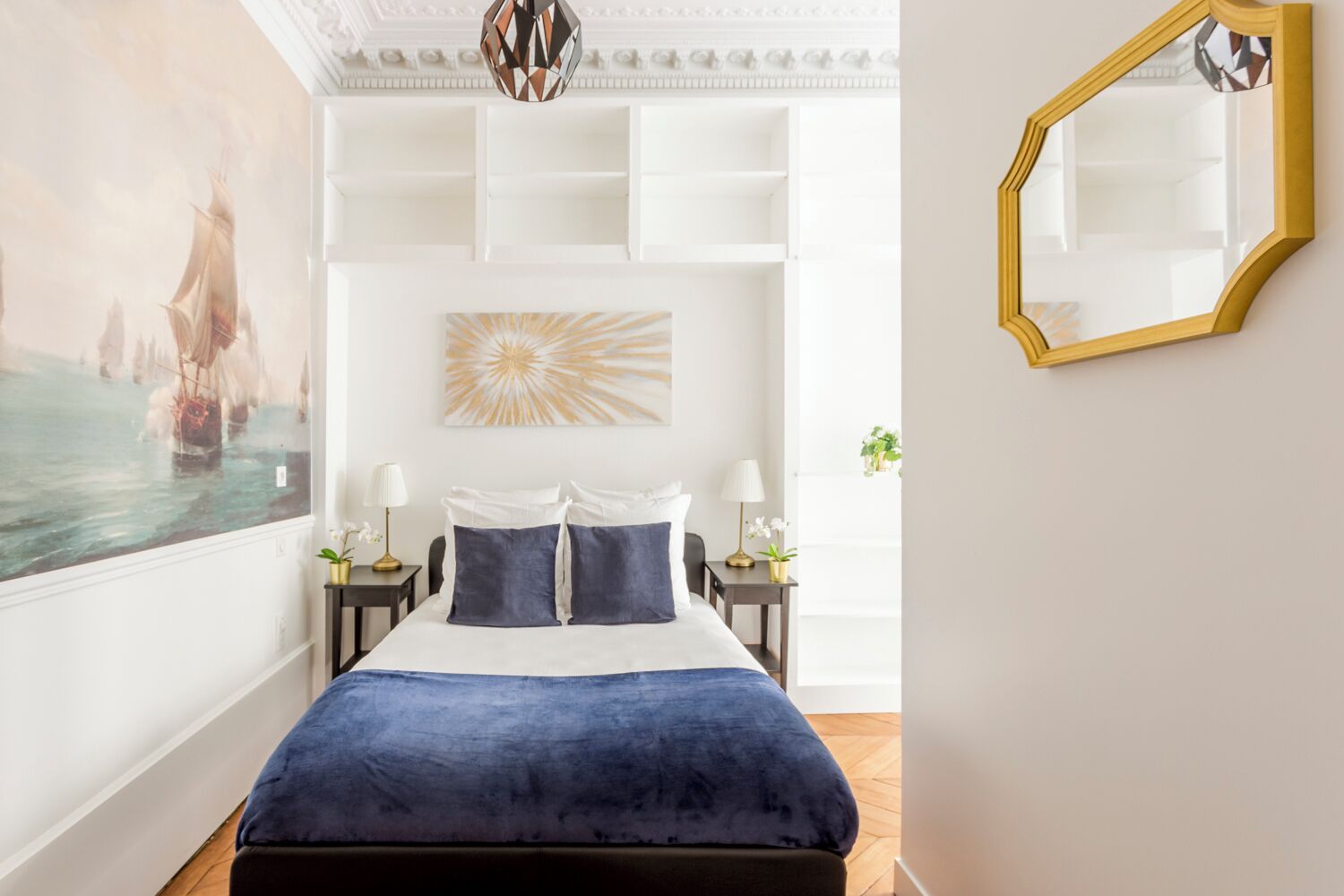 A bedroom with a large bed, two bedside tables, a wall-mounted mirror, and a painting of a ship on the left wall.