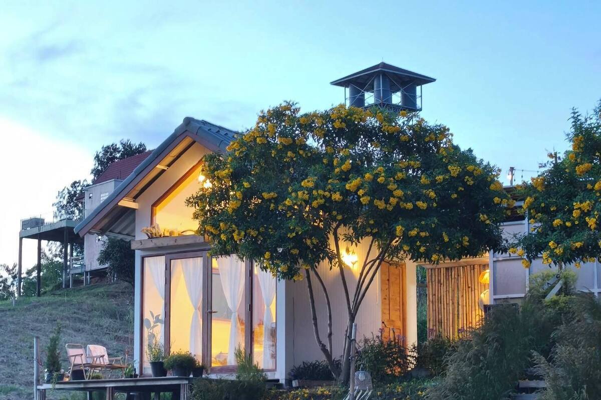 Una casa moderna con un amplio ventanal, una terraza de madera y un árbol con flores amarillas.