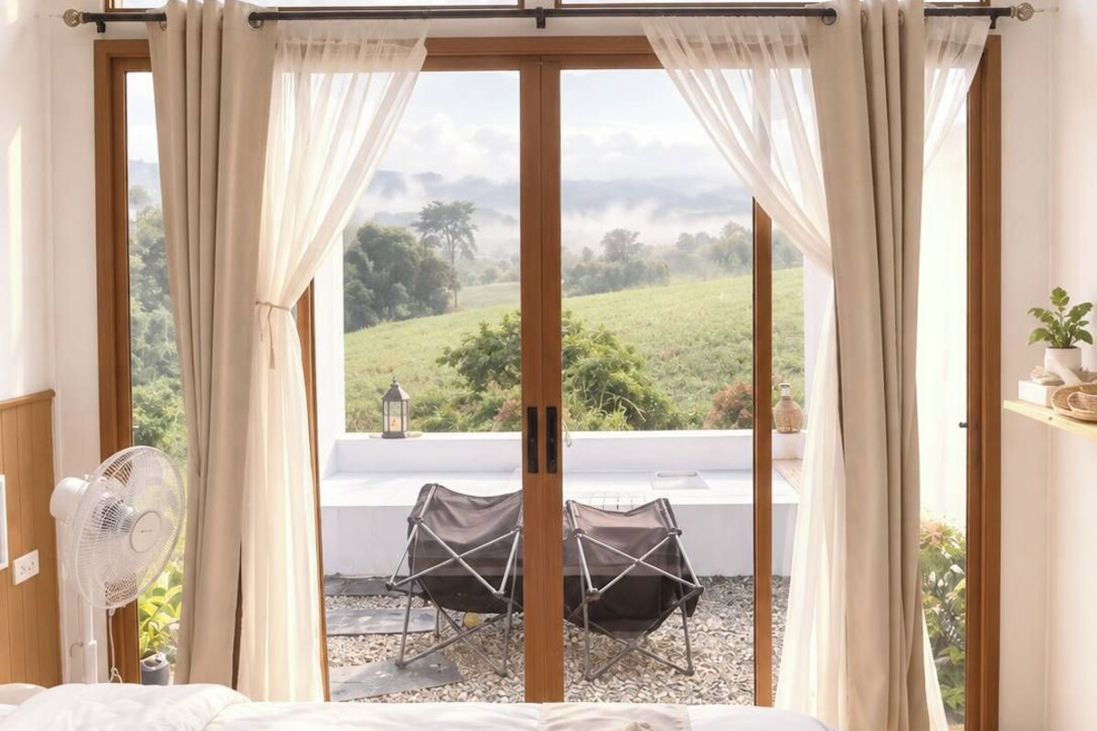 A bedroom with a large bed, a fan, and a view of a green landscape through a glass door.
