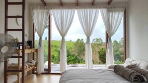 A bedroom with a bed, a fan, a wooden ladder, and a view of greenery outside.
