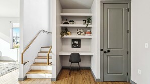 Un interior moderno con una escalera de madera, estantes blancos con plantas y libros, y una puerta gris.