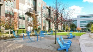 Un área de estar al aire libre con sillas y mesas azules, rodeada por un edificio moderno con ventanas amplias y un césped.