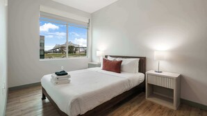 A modern bedroom with a large bed, a nightstand, a ceiling fan, and a window with a view of a building and trees. - Riverscape Townhome (Baton Rouge)