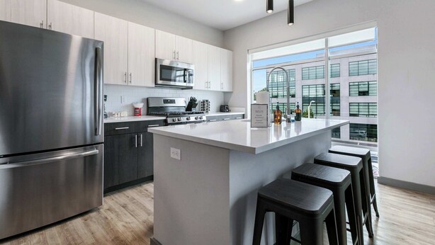 A modern kitchen with stainless steel appliances, a central island with stools, and a large window offering a view of the building's exterior. - Peaceful Modern Townhome | Water Campus (Baton Rouge)