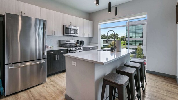 A modern kitchen with stainless steel appliances, a large island with bar stools, and a view of the outside through a large window.