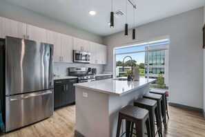 Une cuisine moderne dotée d’appareils électroménagers en acier inoxydable, d’un grand îlot central avec des tabourets, et offrant une vue sur l’extérieur grâce à une grande fenêtre.