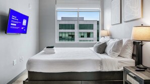 A modern hotel room with a large bed, a flat-screen TV, a window with a city view, and a lamp on a bedside table.
