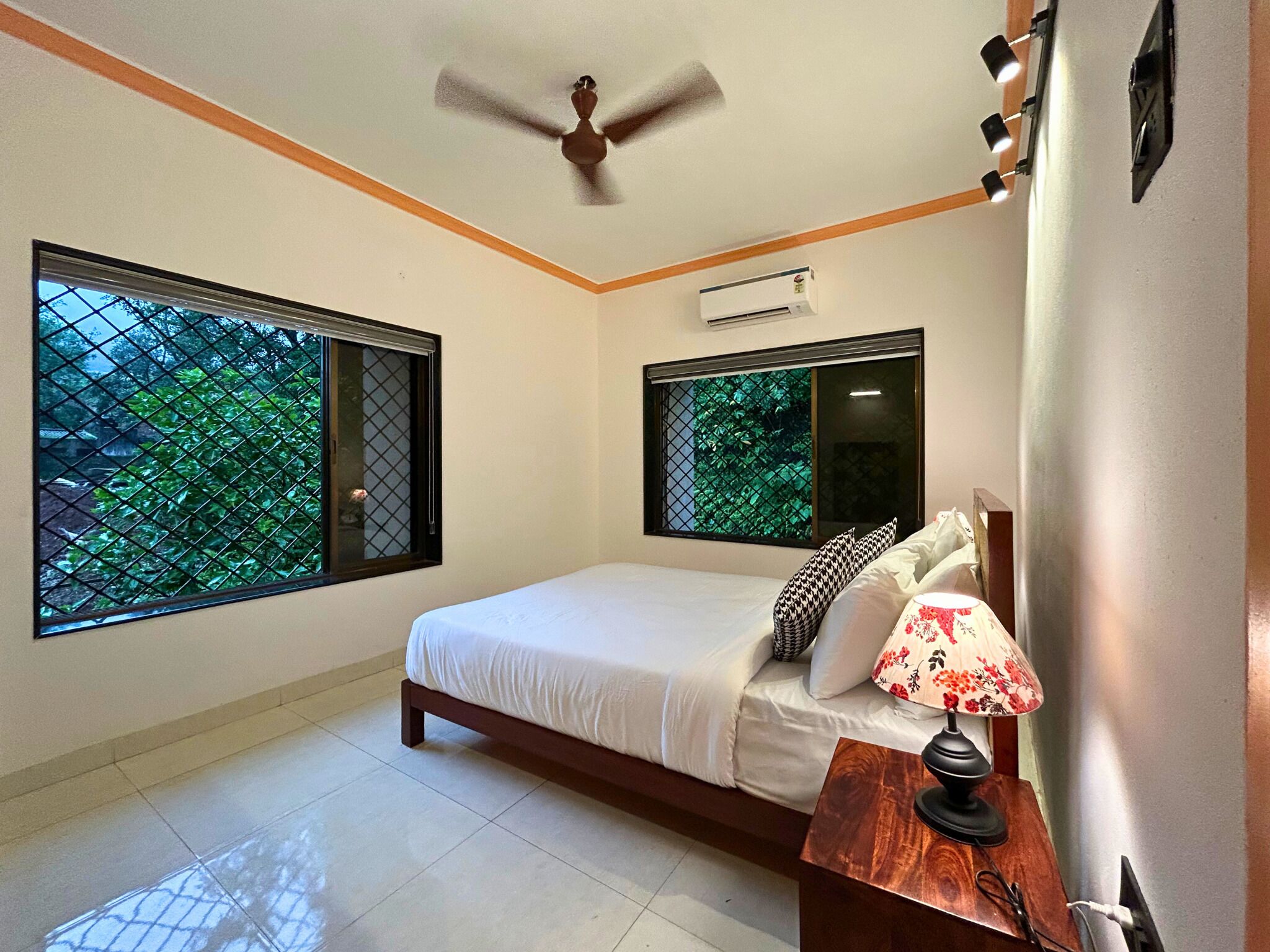 A bedroom with a bed, a ceiling fan, two windows with metal grilles, a bedside table with a lamp, and an air conditioning unit.