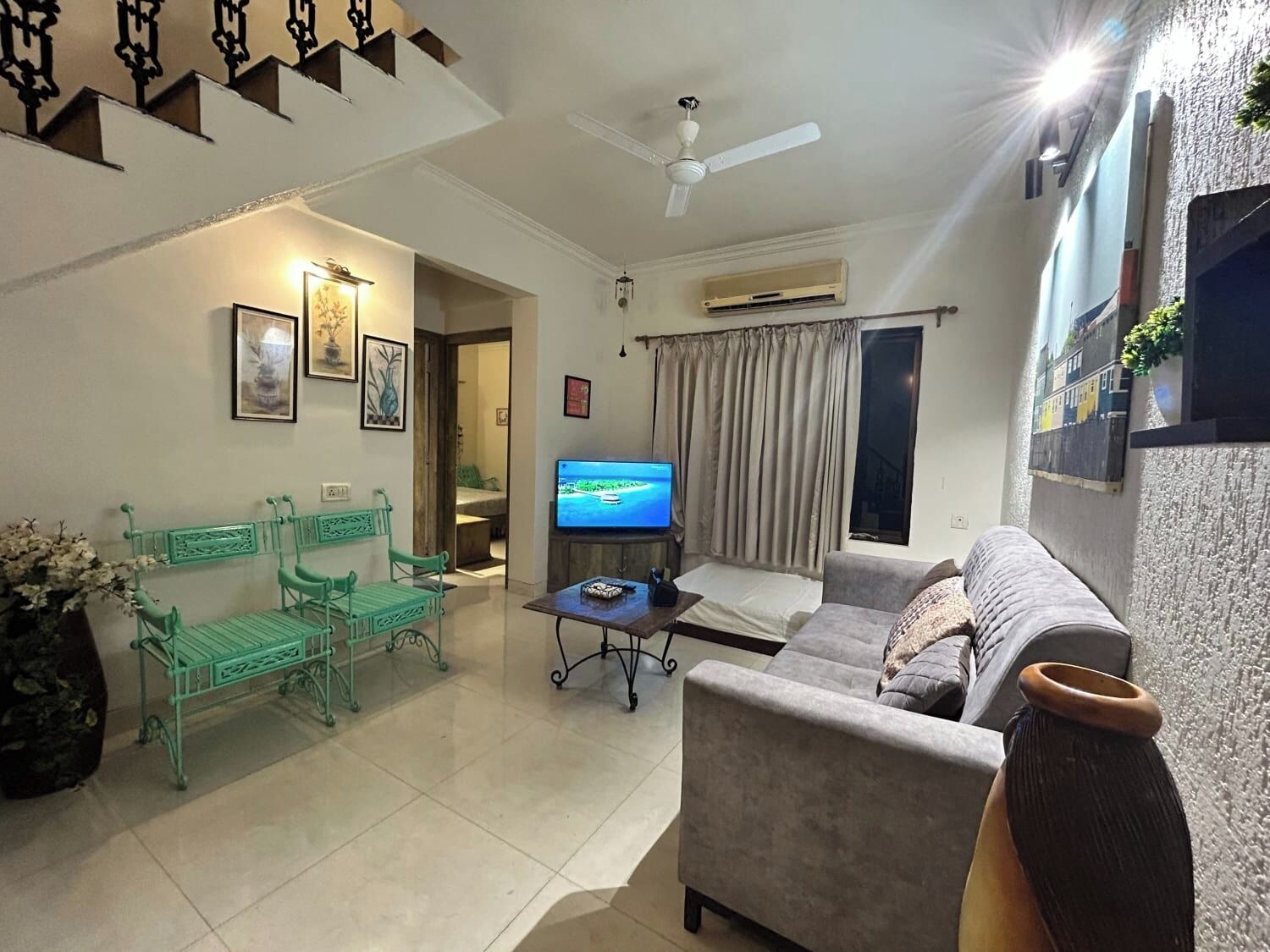 Una sala de estar con un sofá, una mesa de centro y un televisor. Hay dos sillones verdes y una planta en maceta.