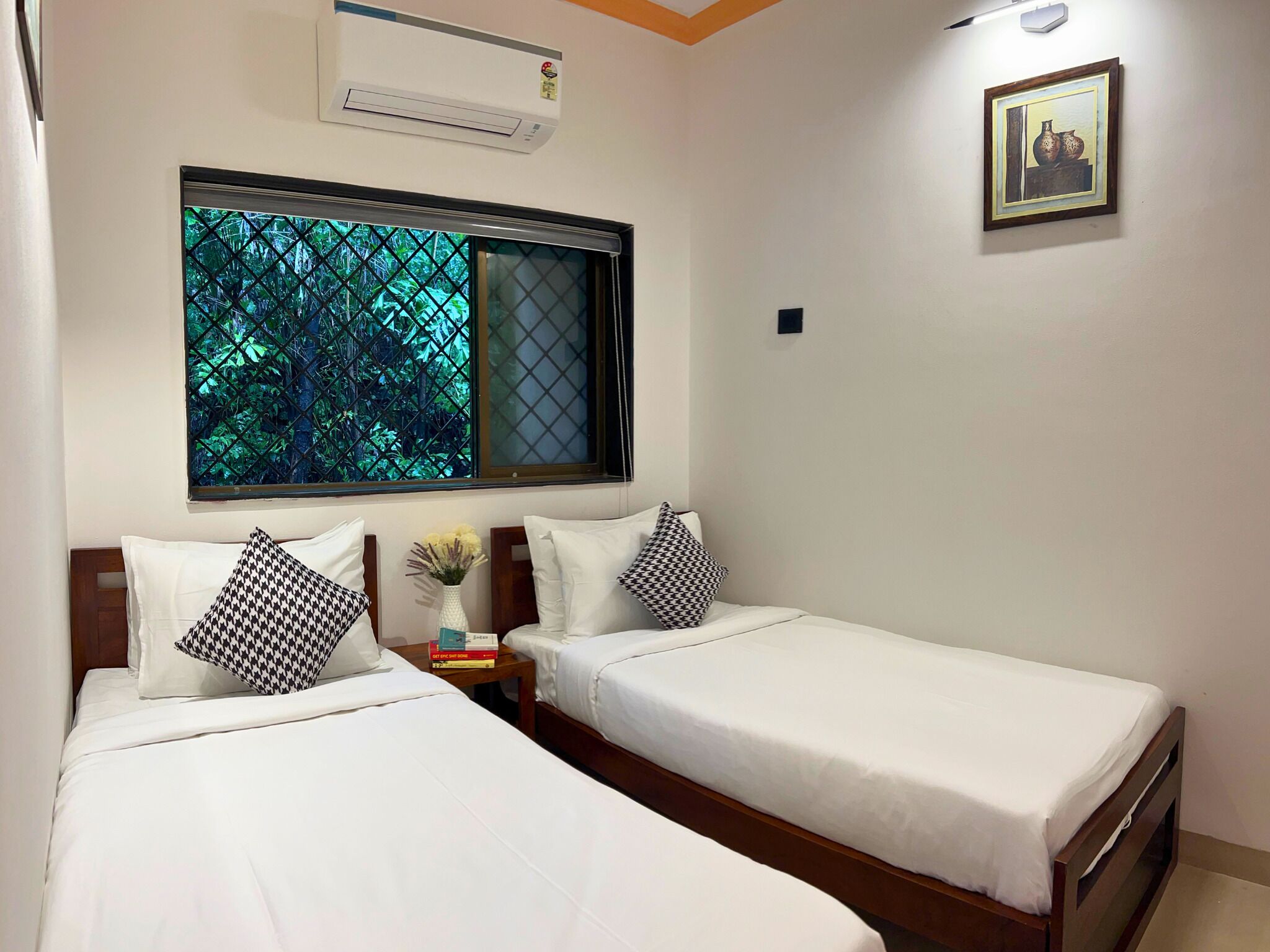 A hotel room with two single beds, a window with a view of greenery, an air conditioner, a small table with a vase, and a framed picture on the wall.