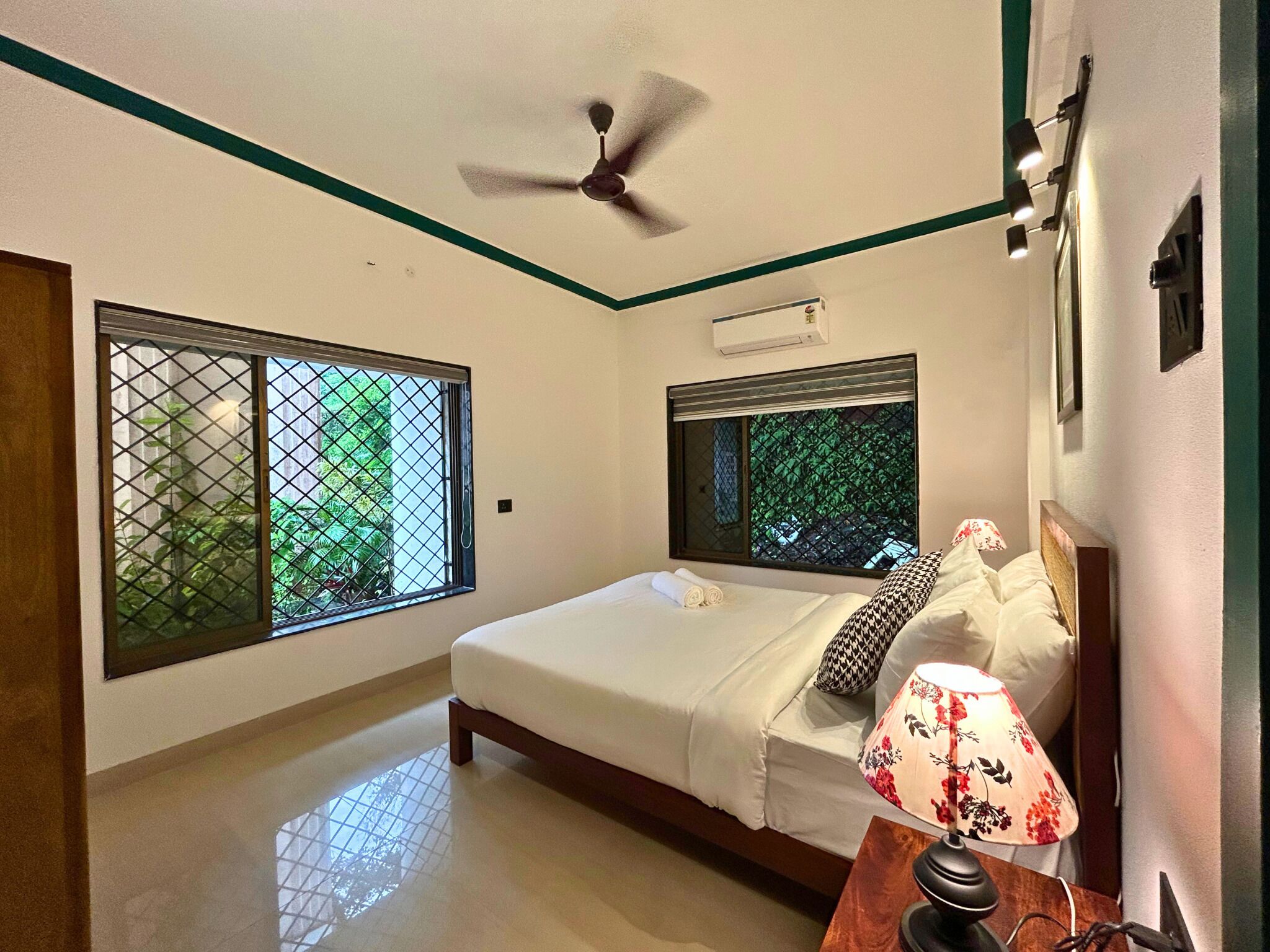 A hotel room with a bed, a ceiling fan, a window with a view of greenery, and a bedside lamp.