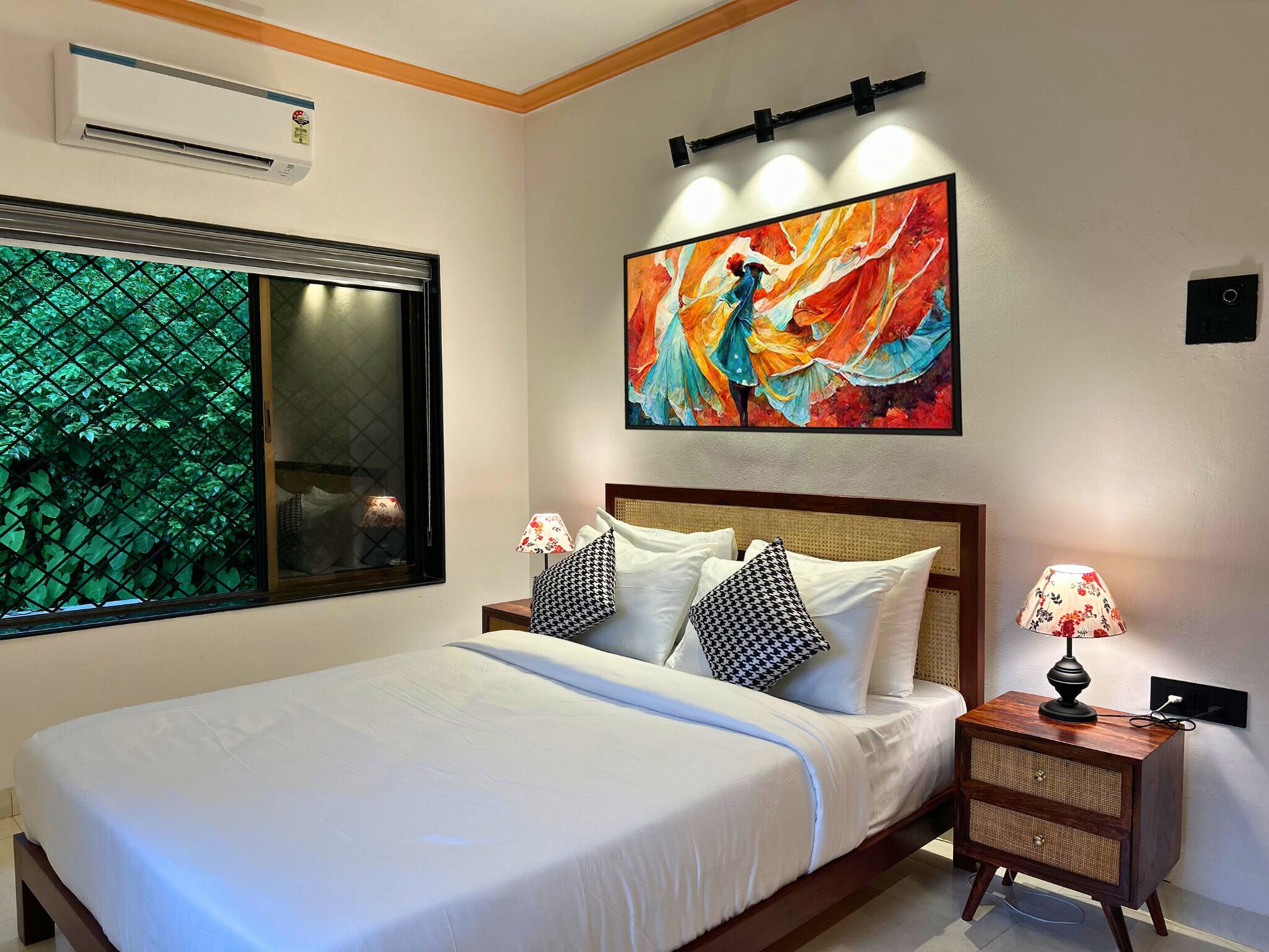 A neatly made bed with white linens, a large window with a view of green foliage, a colorful abstract painting above the bed, and a small bedside table with a lamp.