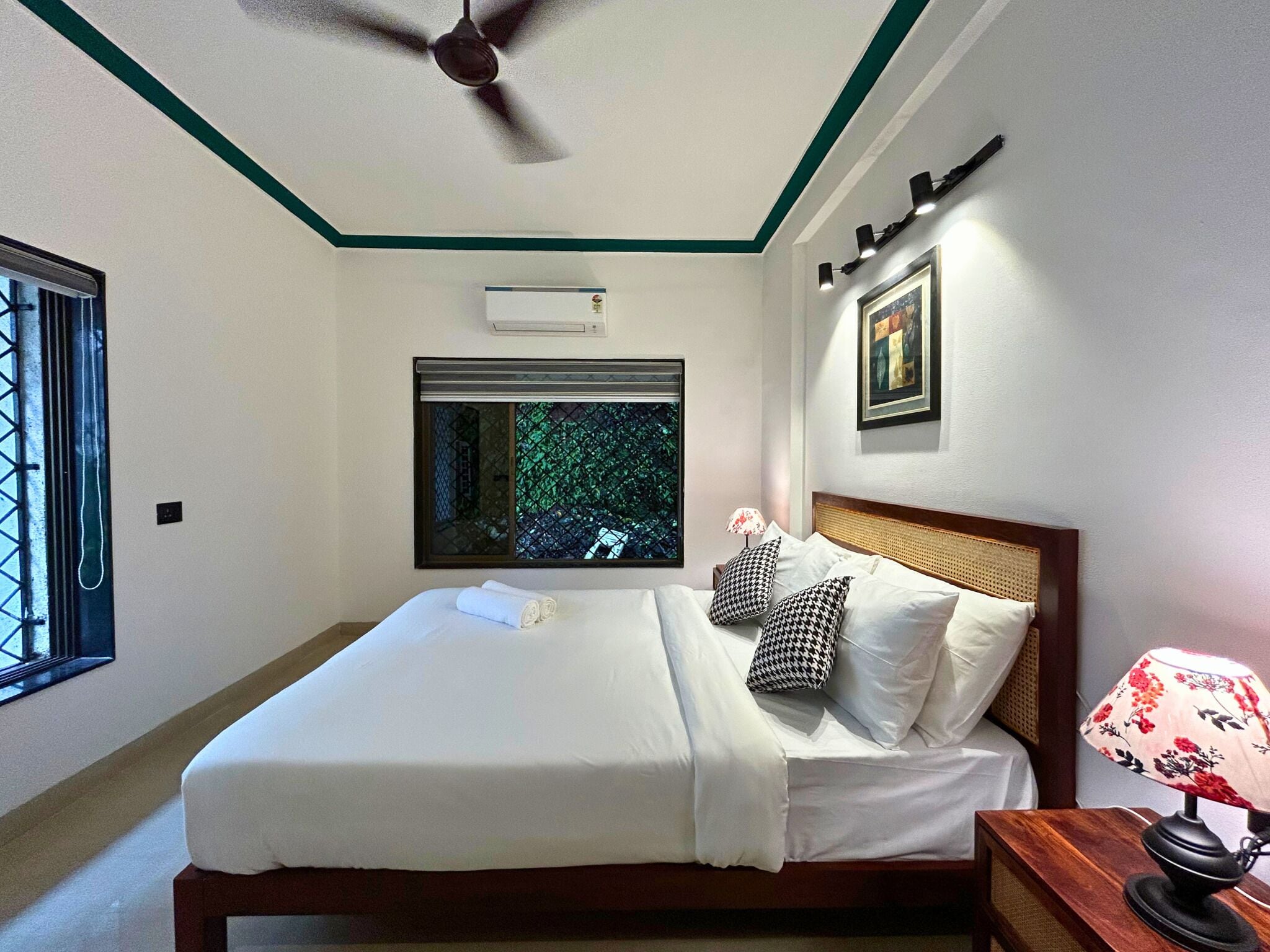 Un lit bien fait, avec du linge de lit blanc, un ventilateur de plafond, une fenêtre donnant sur un paysage verdoyant et un climatiseur fixé au mur.