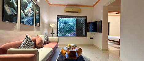Un salón moderno con un sofá, una mesa de centro y una televisión. Hay una ventana con vistas a árboles y un equipo de aire acondicionado en la pared.