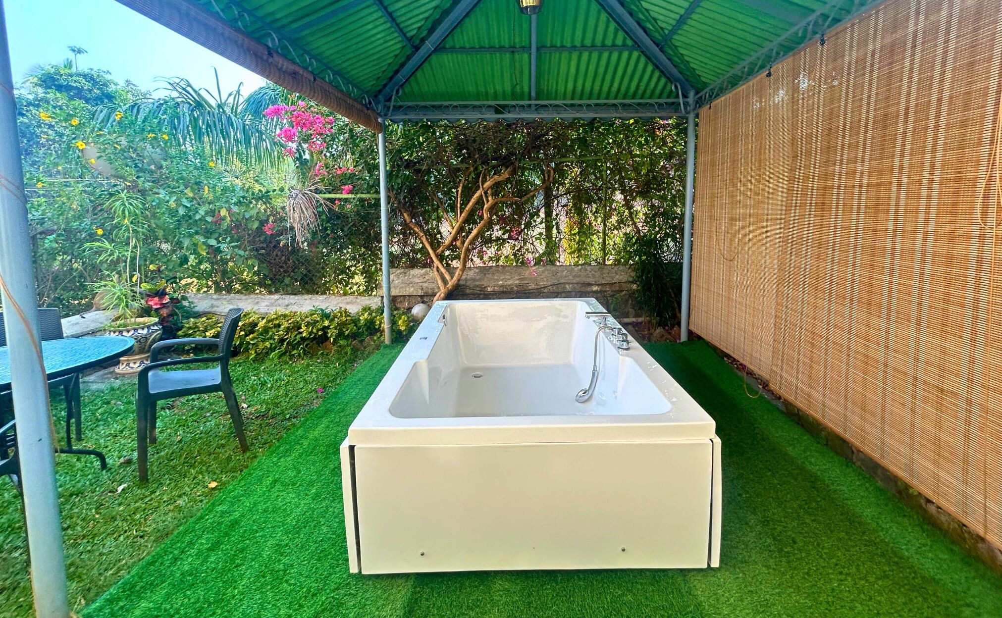 Un área de spa con una bañera blanca grande bajo un techo verde, rodeada de exuberante vegetación y una cerca de bambú.