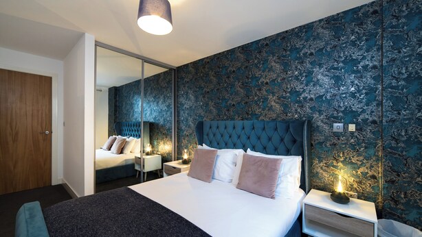 Una habitación de hotel moderna con una cama grande, una mesita de noche con lámpara, un espejo y un armario.