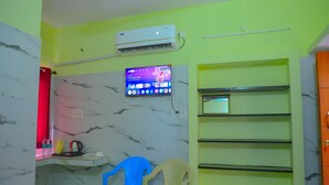 A room with a marble-patterned wall, a mounted air conditioner, a flat-screen TV, a shelf unit, and a desk with a chair. - RR RESIDENCY RANIPET (Walajapet)