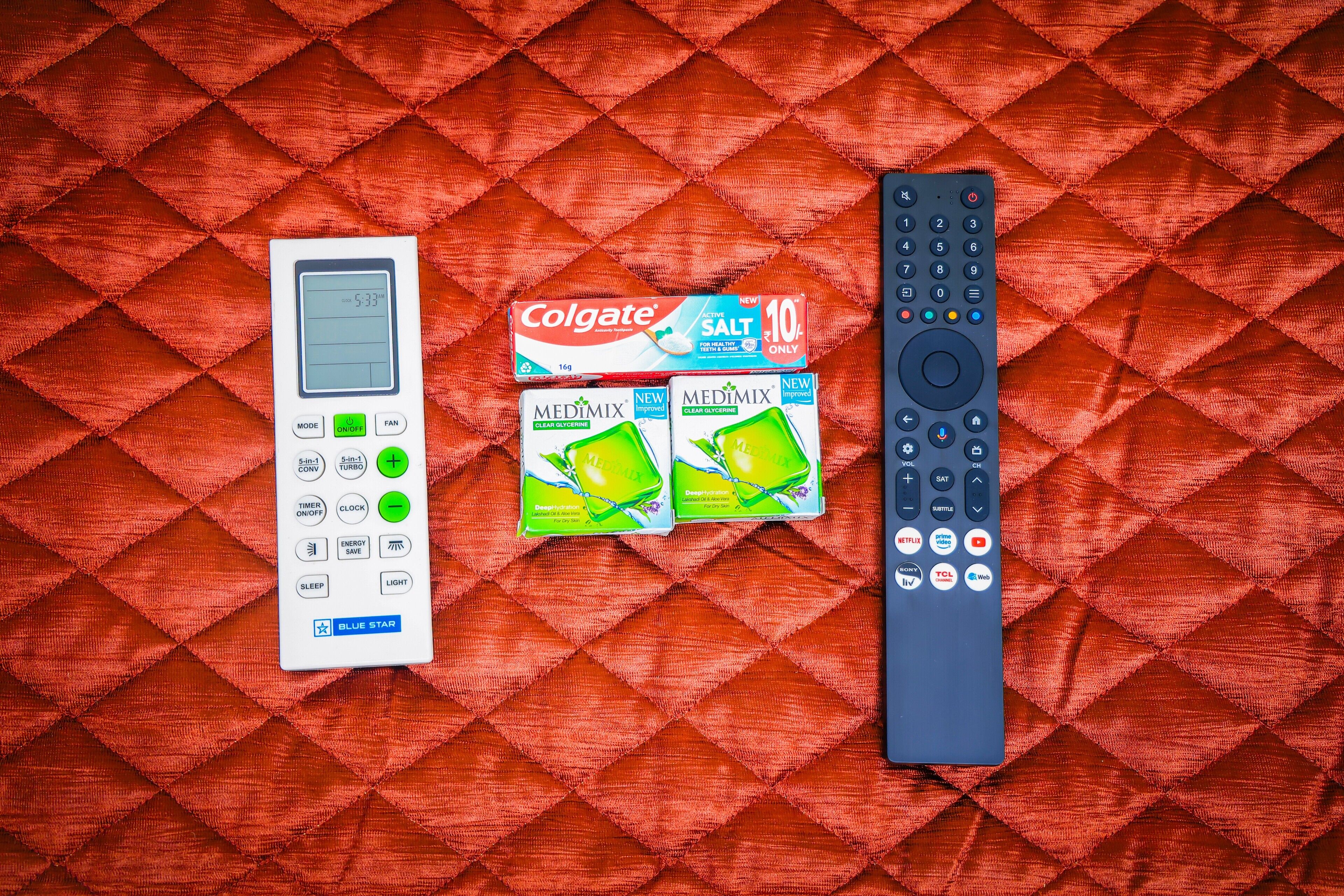 Et hvidt digitalt ur, en Colgate tandpastaæske og en fjernbetjening på en rød, quiltet overflade.