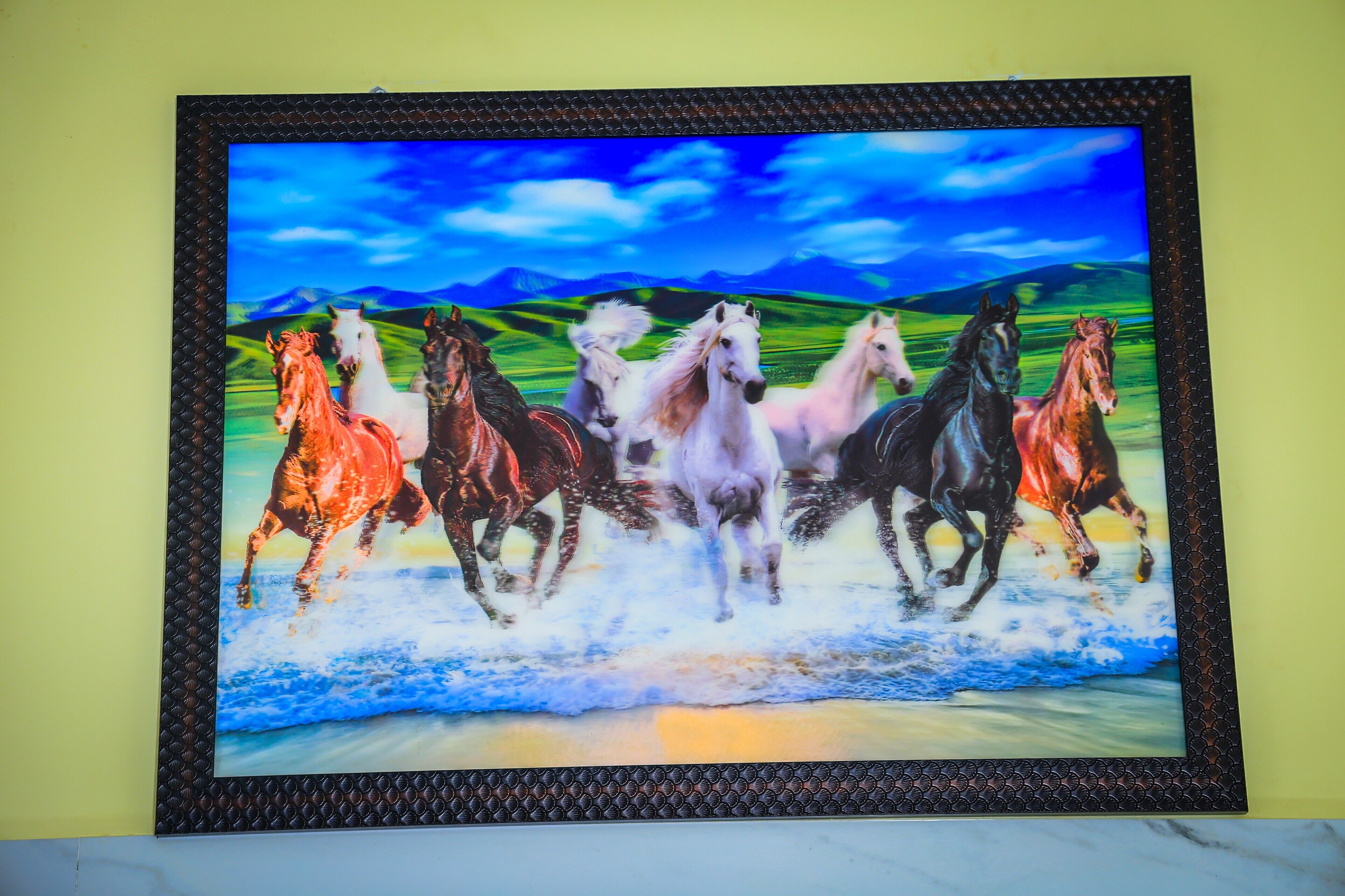 Una obra de arte enmarco que muestra caballos corriendo en una playa, con montañas al fondo.
