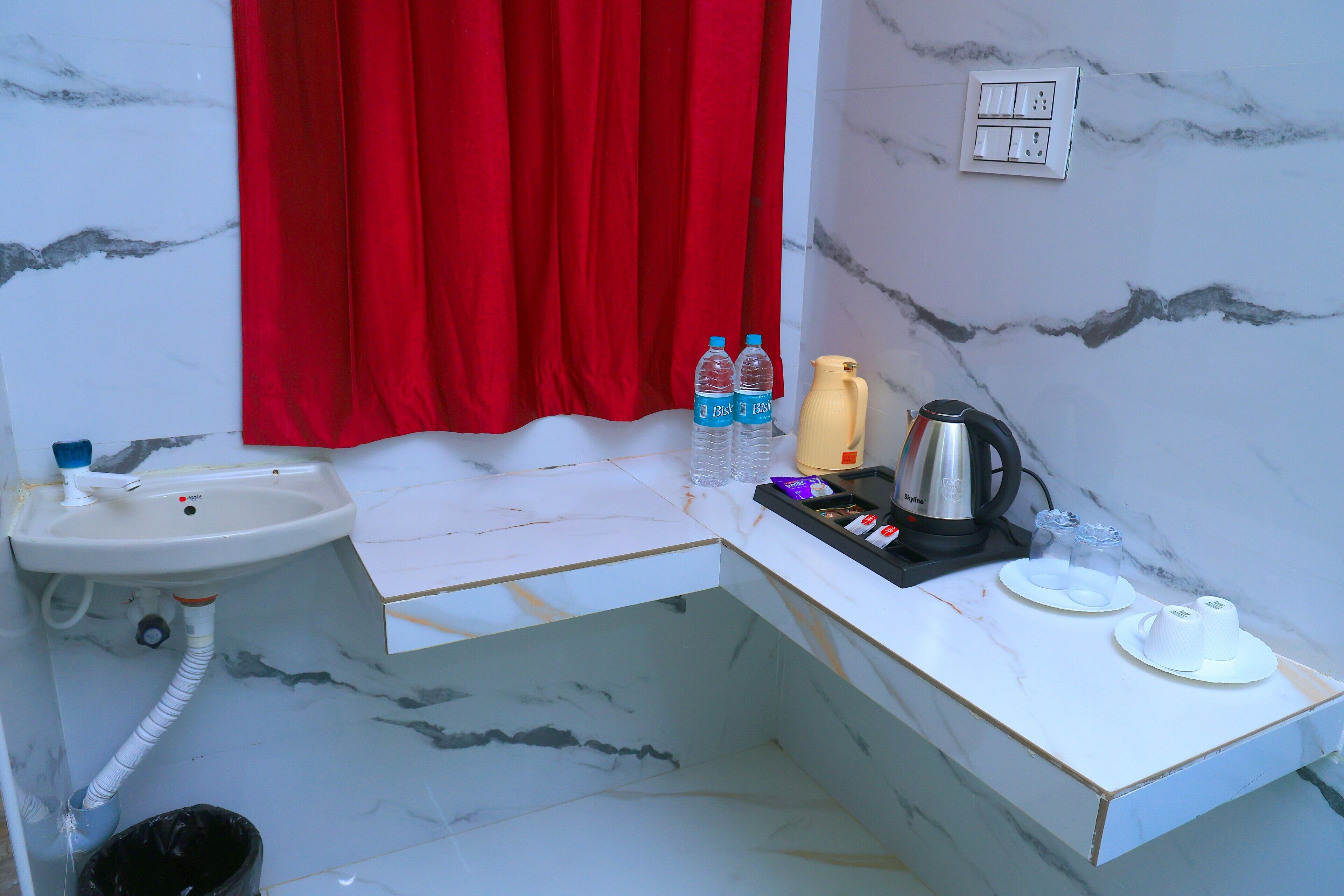 Une salle de bain avec un lavabo, un rideau rouge, deux bouteilles d’eau, une bouteille jaune, une bouilloire et une assiette avec des tasses.